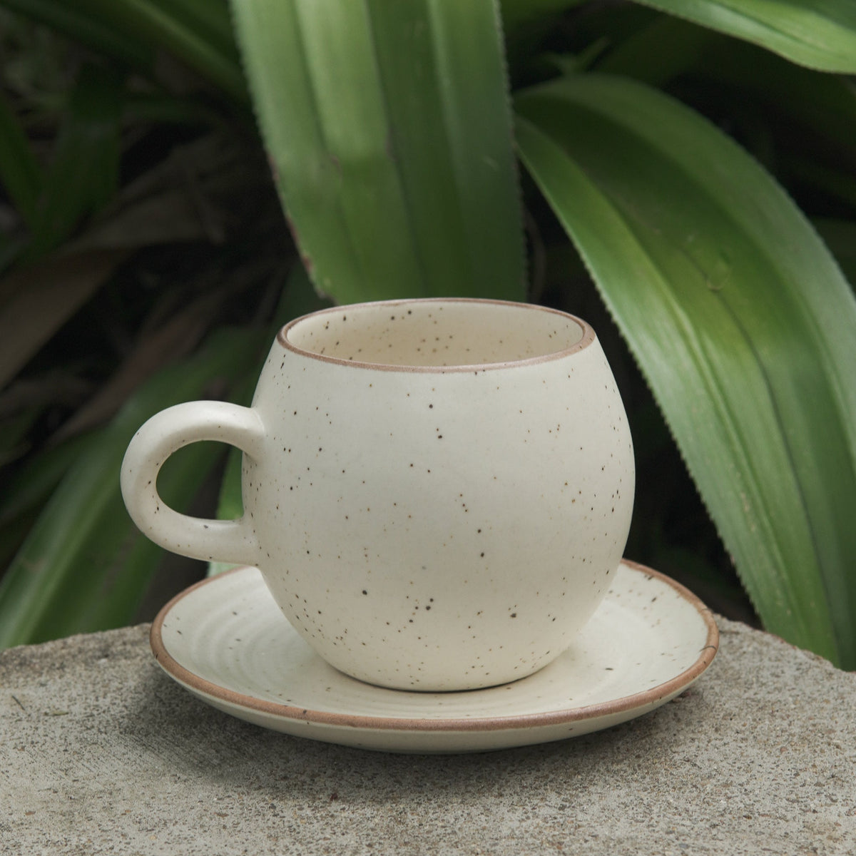 A beige stoneware bubble mug with a matching saucer, placed on a stone surface with green foliage in the background.