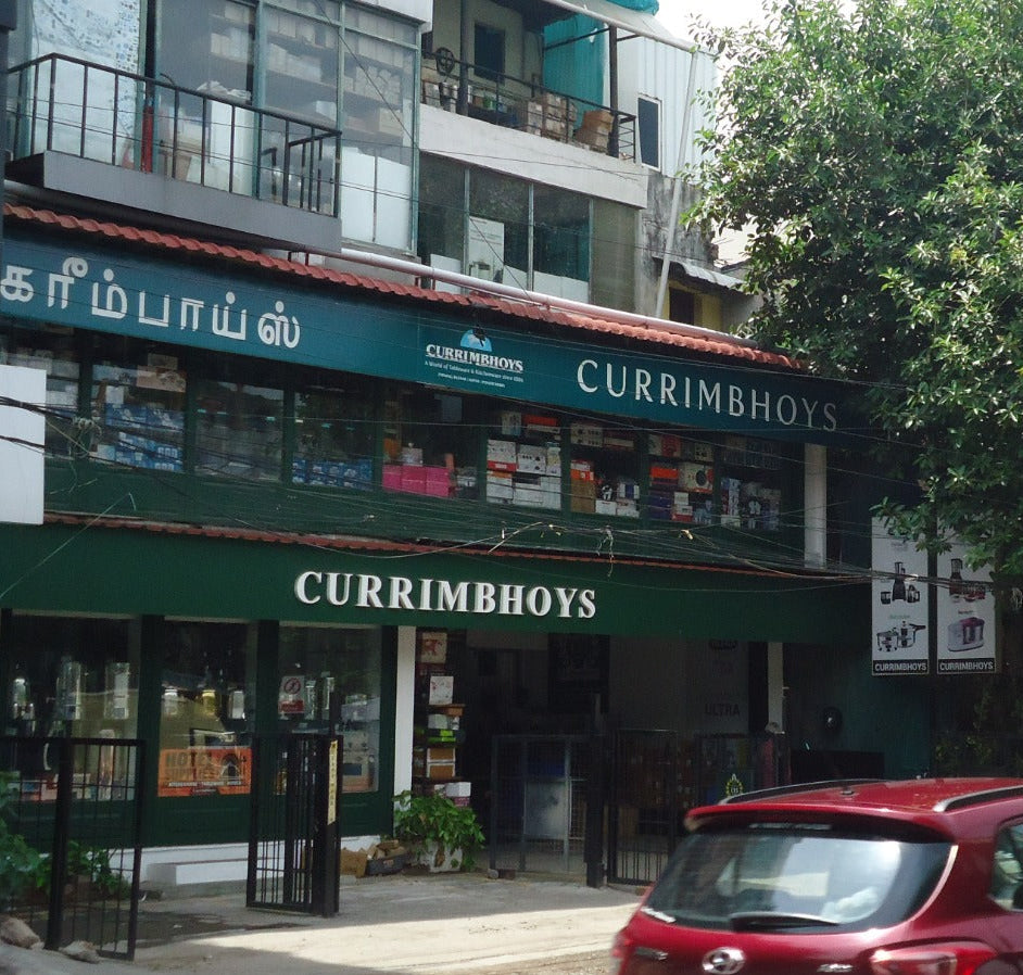 A street view of the Currimhoys store in Chennai, featuring a dark green storefront with white lettering, a red car in the foreground, and a large green tree on the right side of the frame.