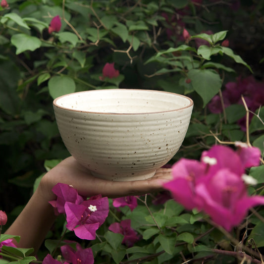 A hand holding a textured stoneware bowl with a plant background featuring pink flowers.