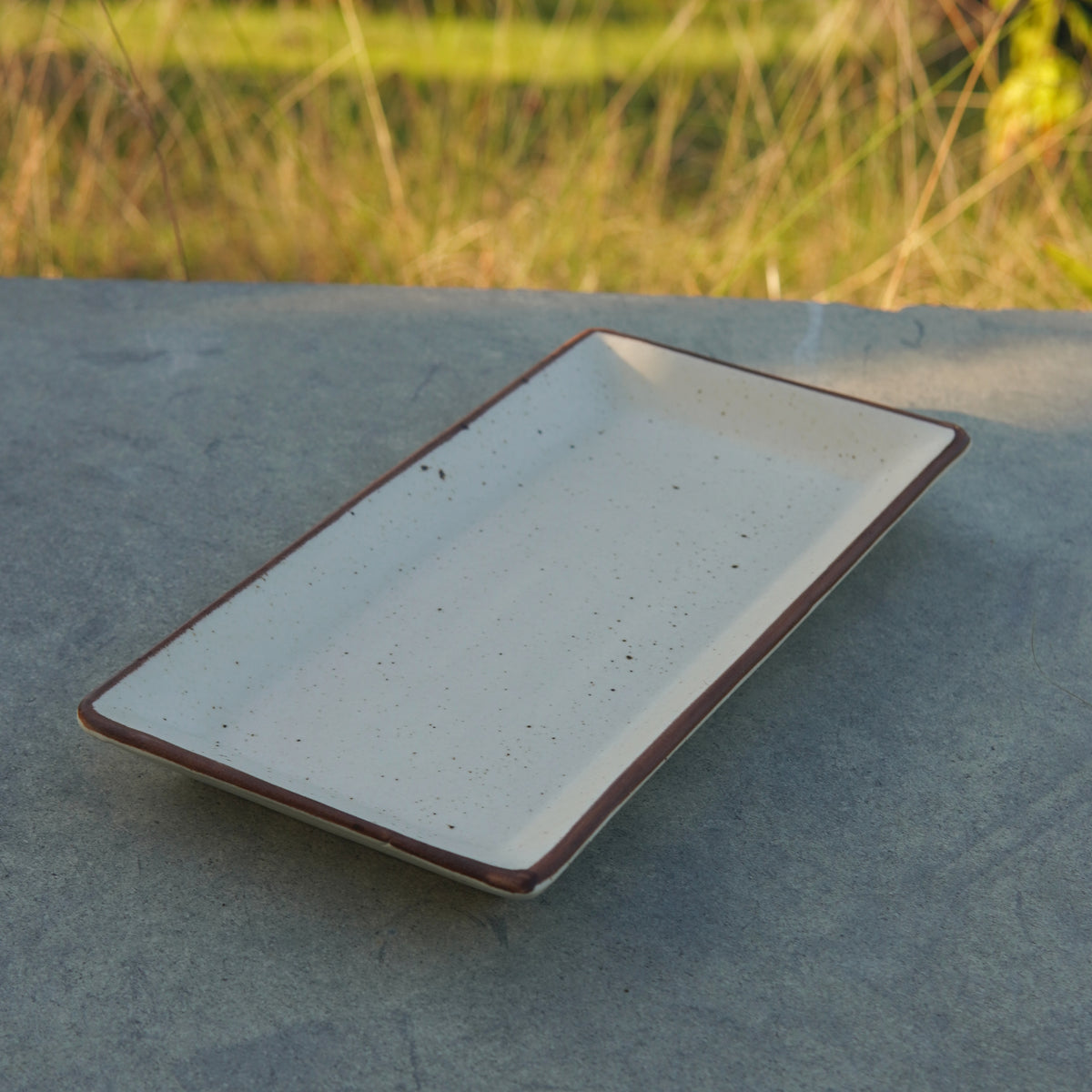 A rectangular stoneware platter with a flat surface and slightly raised edges, displayed on an outdoor table with a natural background.