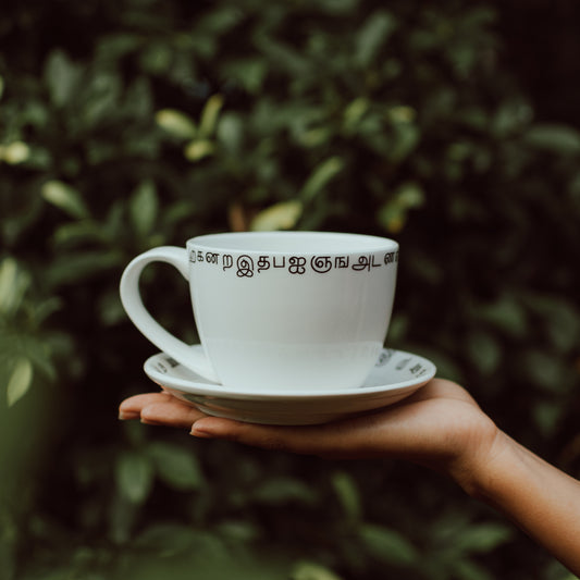 A hand holding a white ceramic coffee cup with black Tamil script design, accompanied by a matching saucer, against a leafy background.